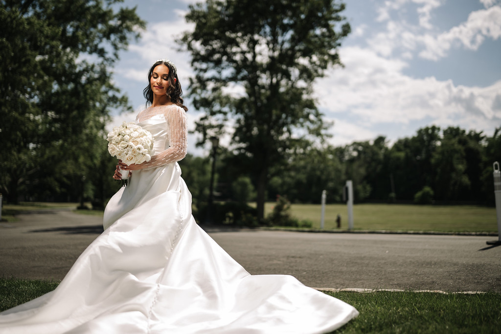 Bride holding a white floral bouquet outdoors, wearing a long sleeve wedding dress with her veil and hair styled neatly, standing on a paved path with trees and sky in the background.