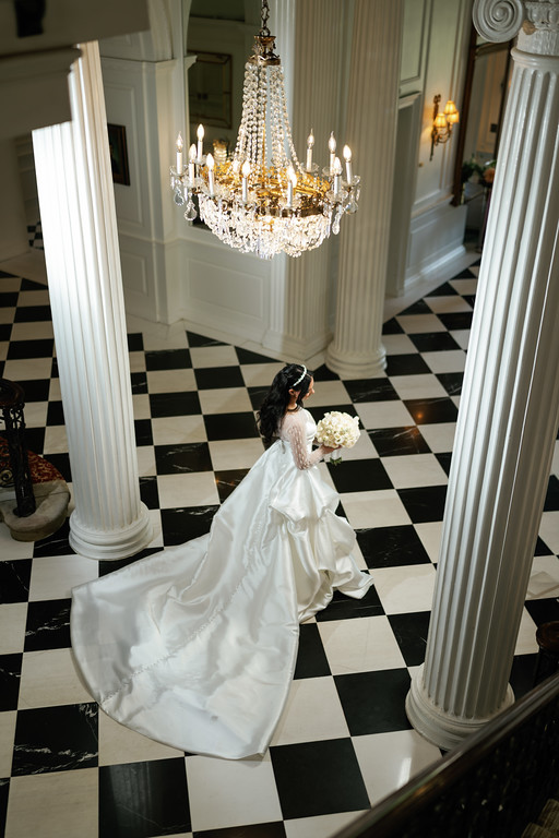 Bride in a white wedding gown holding a bouquet, standing in a hall between tall columns with a large chandelier above.