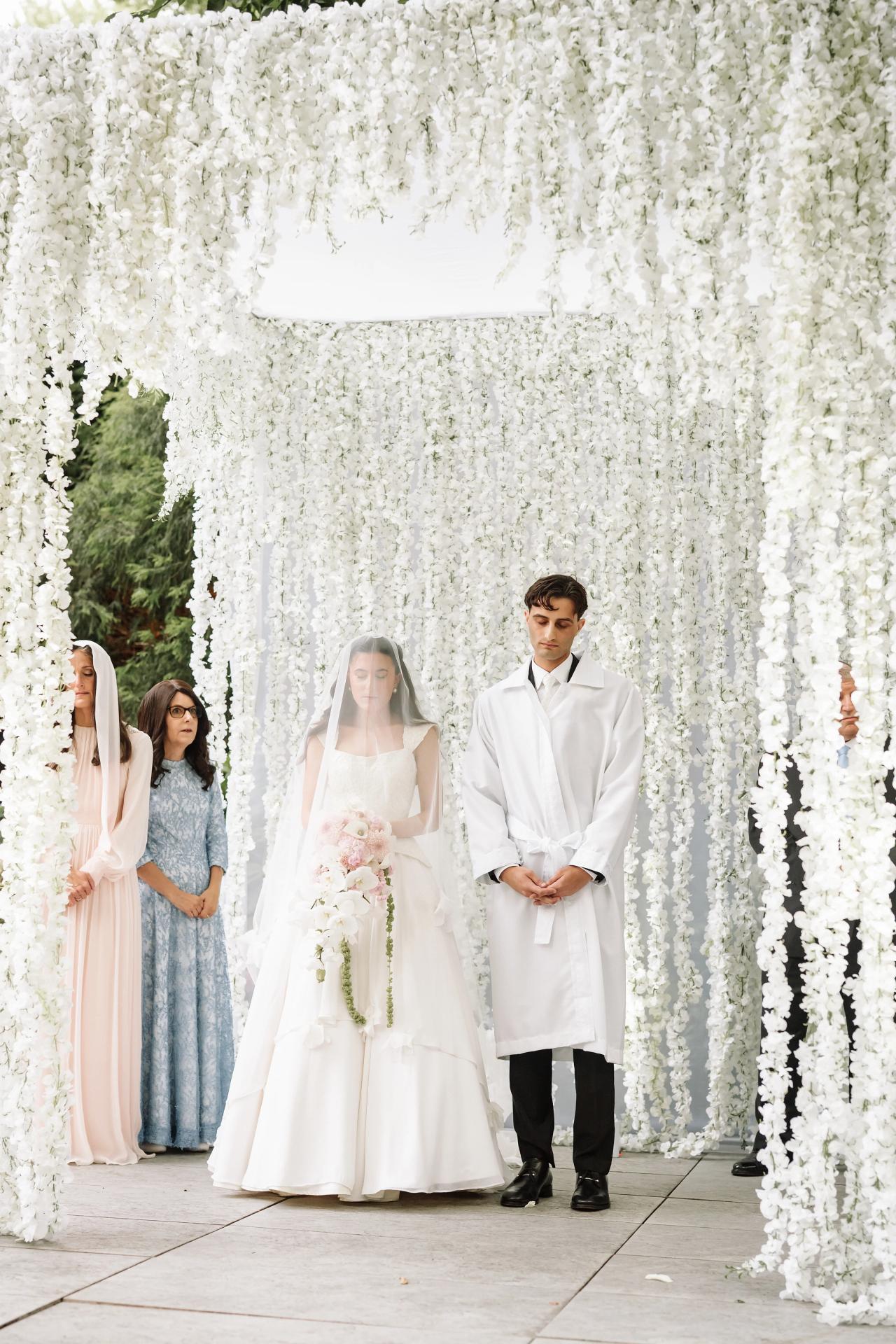Bride and groom standing under an ornate white floral chuppah with close family members beside them during a wedding ceremony outdoors. Wedding photography at Crystal Plaza, Livingston, NJ.