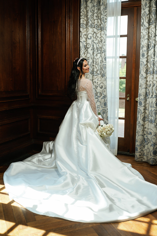 Bride in a white wedding gown seated indoors near a wooden door and window, holding a bouquet in hand as sunlight falls across her dress.