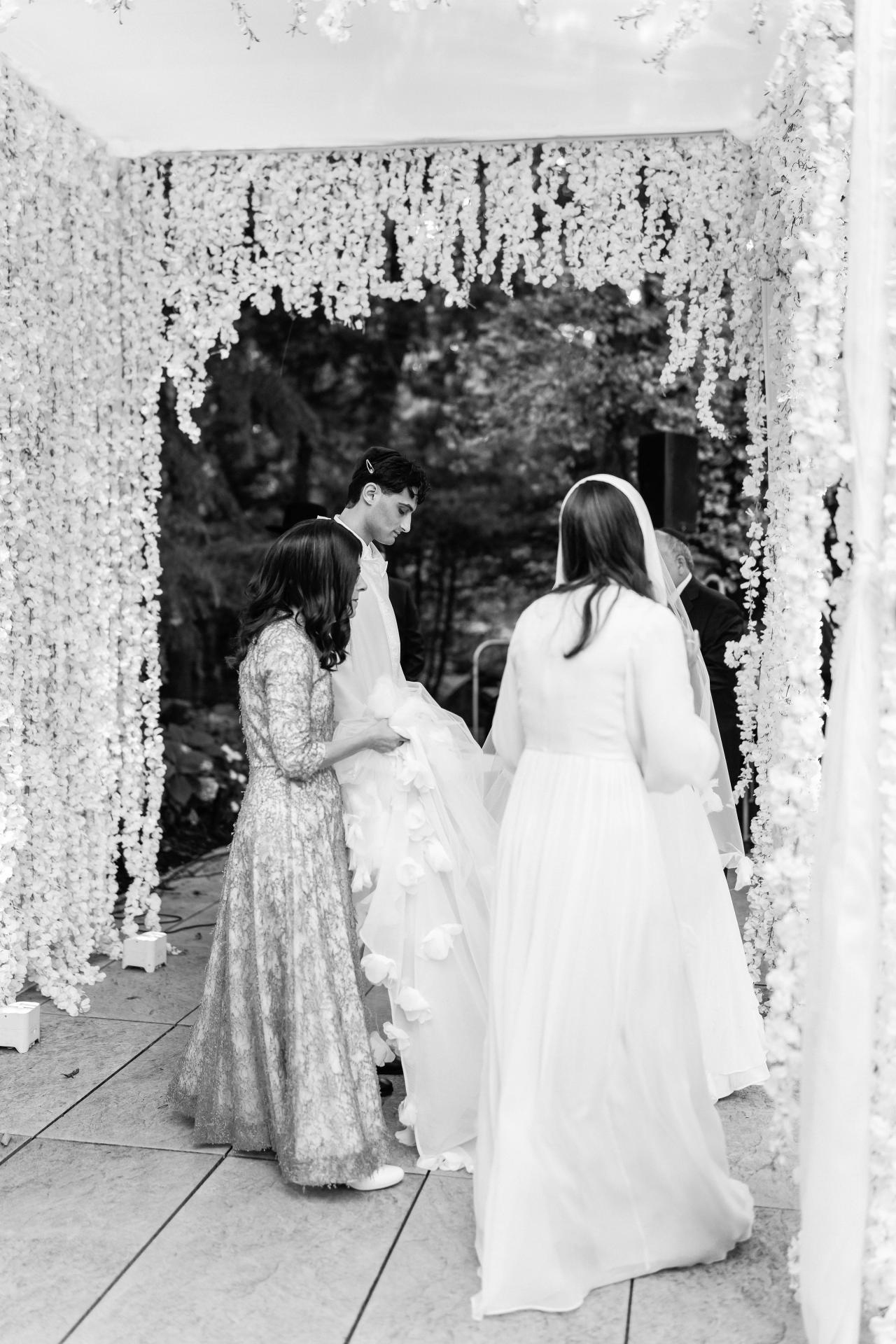 Black-and-white photo of a bride and groom sharing an intimate moment during the badeken, framed by floral garlands and draped fabric at an outdoor ceremony. Wedding photography at Crystal Plaza, Livingston, NJ.