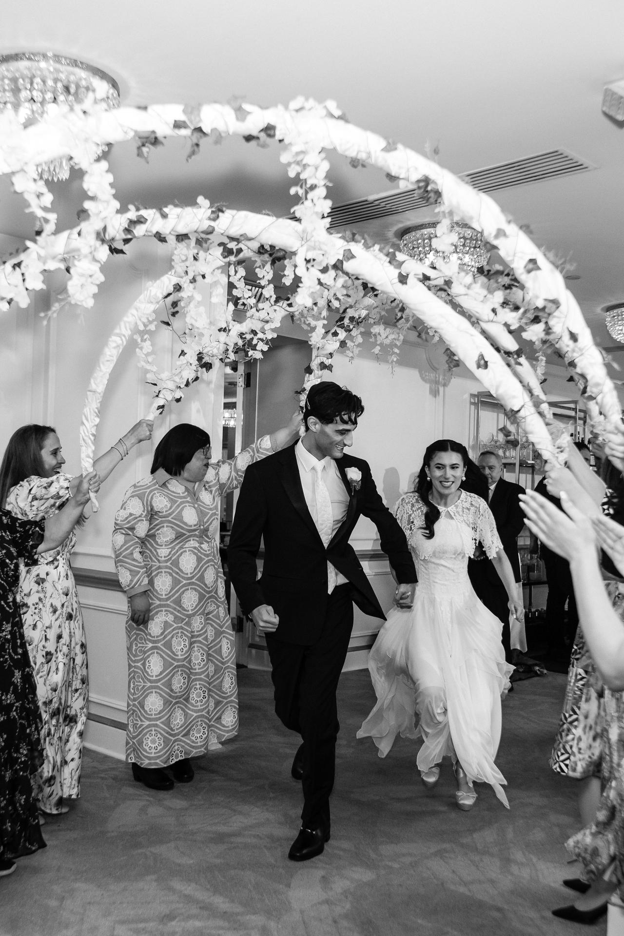 Black-and-white wedding dance moment: bride and groom walking forward under a chuppah with guests cheering and throwing confetti as hands reach toward them.