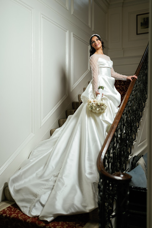 Bride in a white ballgown holding a bouquet while standing on a staircase landing, smiling toward the camera in an elegant indoor setting.
