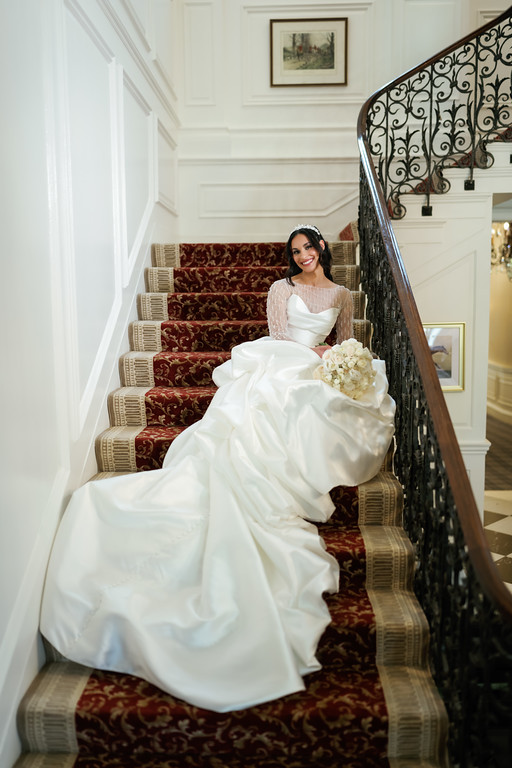 Bride in a white wedding gown seated on an indoor staircase, smiling and holding her bouquet, framed by ornate railings and bright walls.