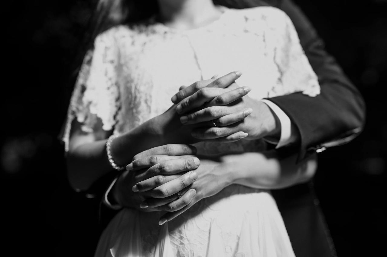Black-and-white close-up of a bride and groom holding hands, lace sleeves visible, fingers intertwined, intimate wedding moment. Wedding photography at Crystal Plaza, Livingston, NJ.