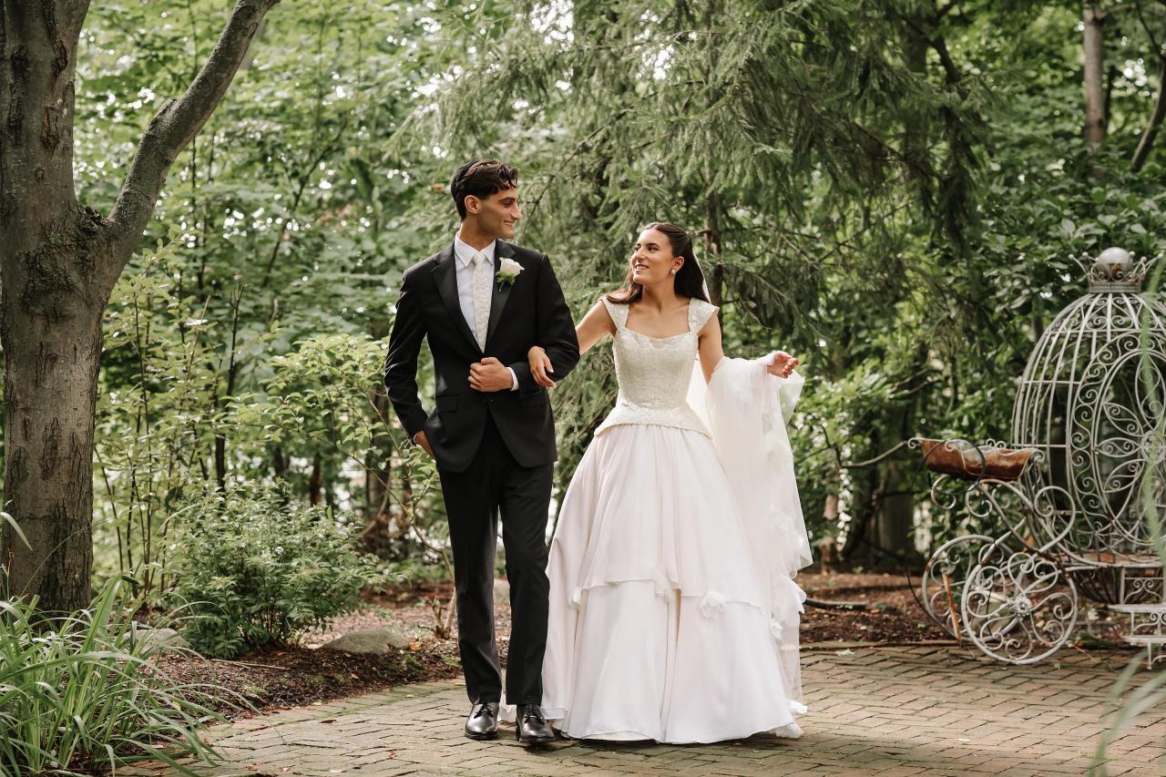 Outdoor garden wedding portrait of a bride and groom walking together along a paved path, smiling and holding hands near lush green trees and decorative metal furniture. Wedding photography at Crystal Plaza, Livingston, NJ.