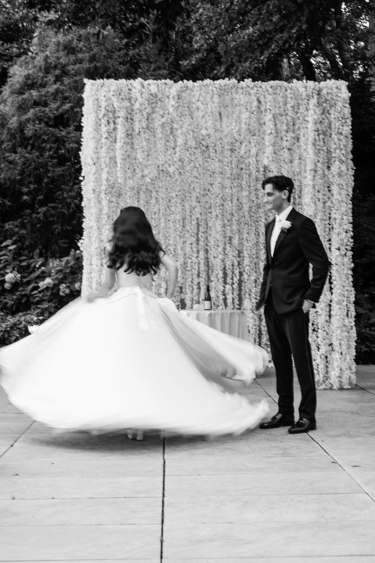 Black-and-white photo of a bride’s flowing wedding gown and veil as she moves in front of a groom standing beside a floral backdrop, outdoors. Wedding photography at Crystal Plaza, Livingston, NJ.