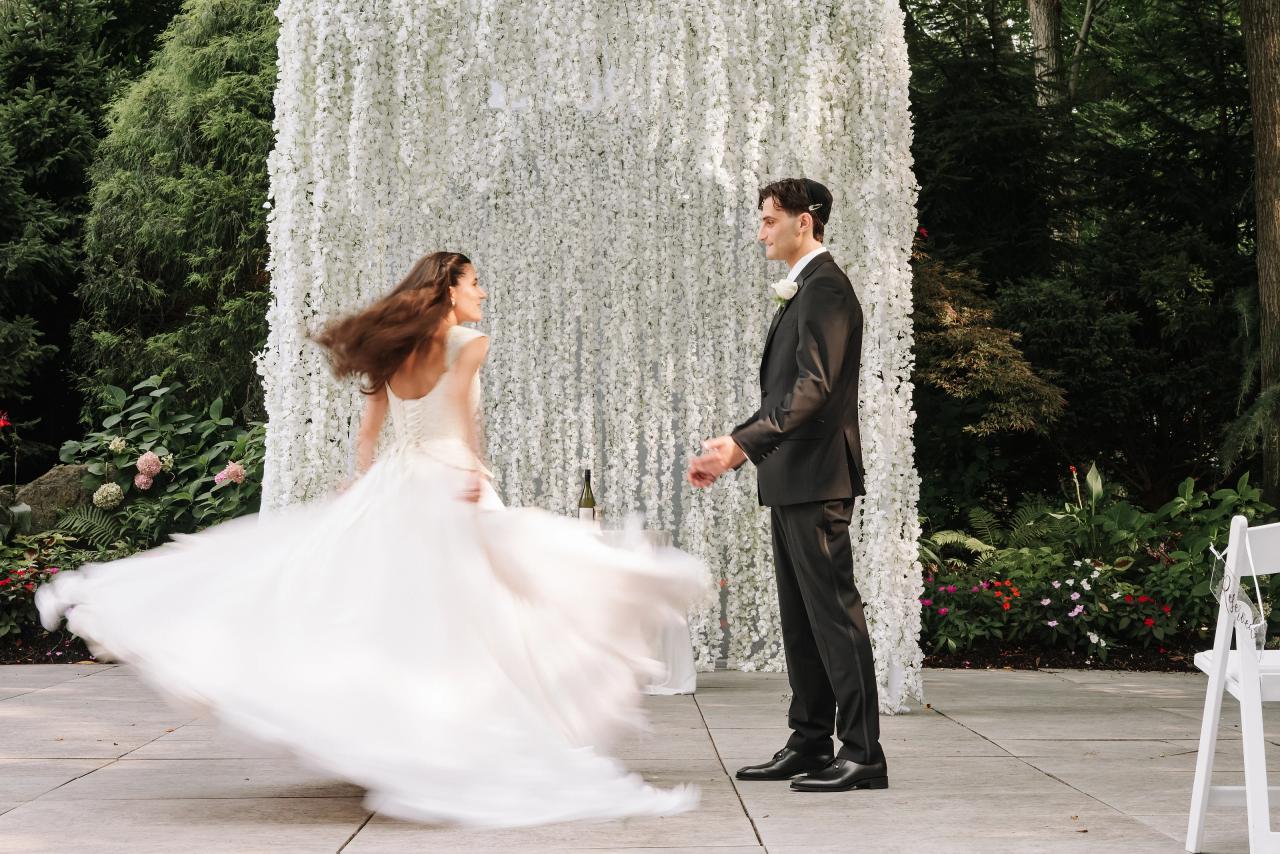Bride and groom in motion near a white floral chuppah backdrop outdoors, with the bride’s dress sweeping across the frame in bright daylight. Wedding photography at Crystal Plaza, Livingston, NJ.