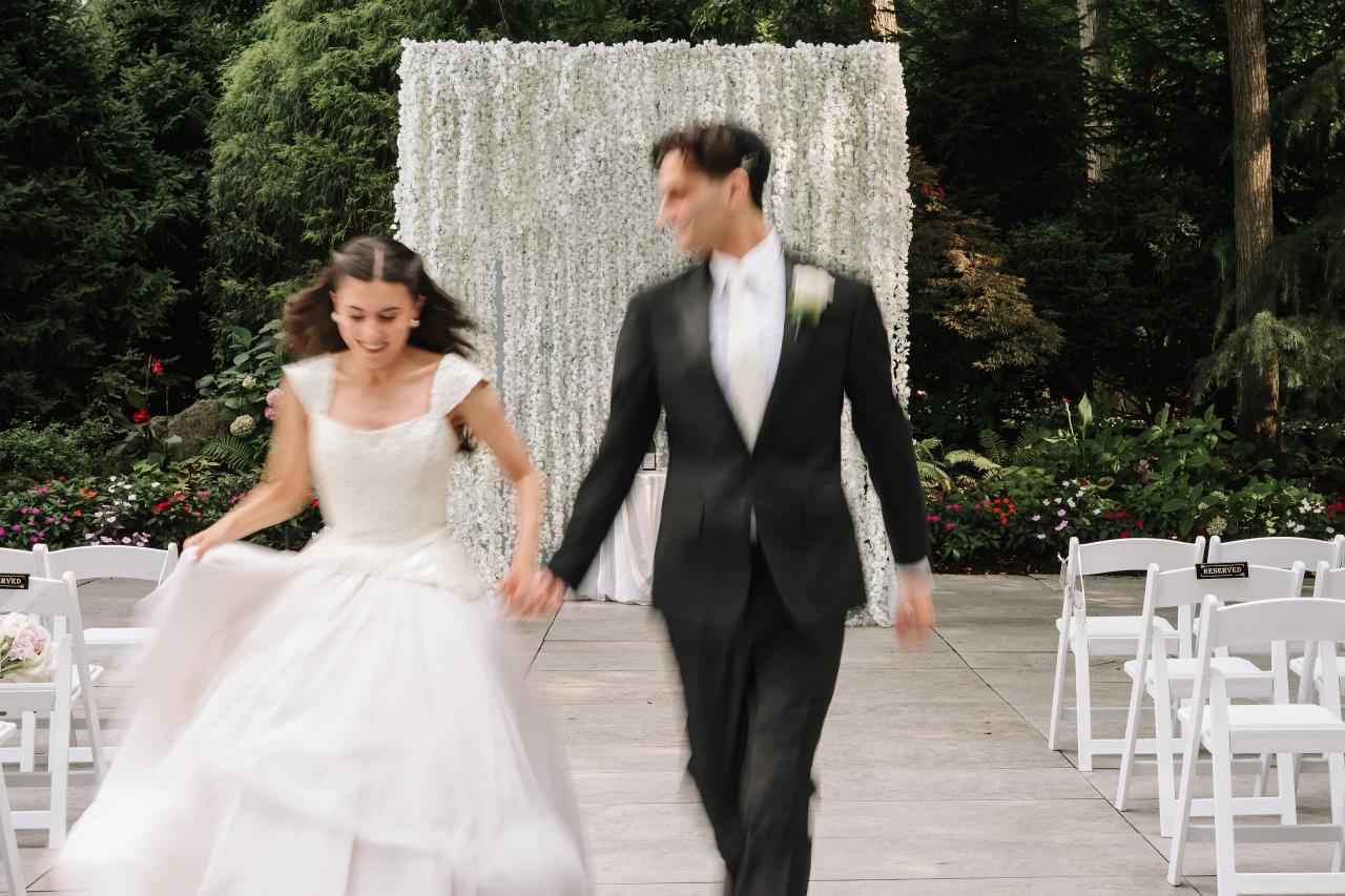 Bride and groom walking together down the aisle outdoors, greenery backdrop and floral arrangements behind them, chairs along both sides, joyful motion. Wedding photography at Crystal Plaza, Livingston, NJ.