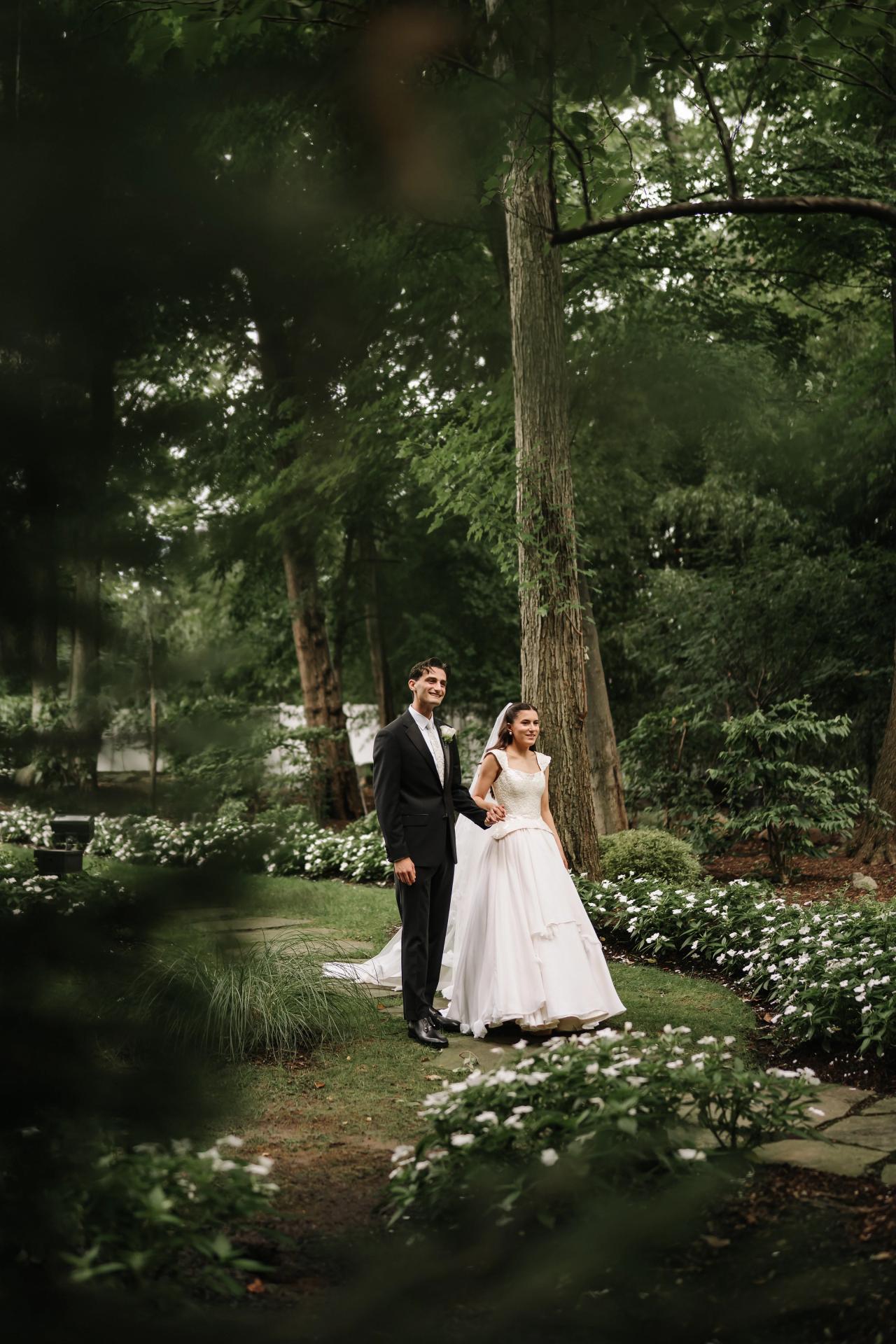 Wedding couple walking together in a shaded garden, surrounded by trees and landscaped flowerbeds. Wedding photography at Crystal Plaza, Livingston, NJ.
