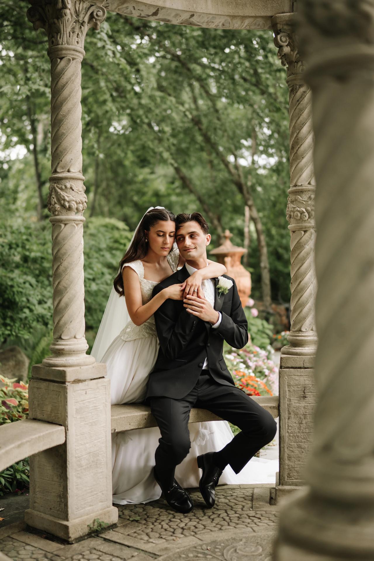 Bride and groom embracing under stone arches in a lush garden, bride seated on a ledge and groom holding her close. Wedding photography at Crystal Plaza, Livingston, NJ.