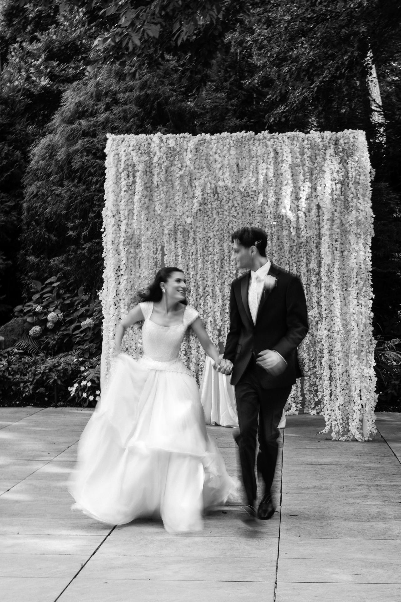 Black-and-white portrait of a bride and groom walking hand-in-hand toward the camera in front of a floral backdrop, captured mid-step. Wedding photography at Crystal Plaza, Livingston, NJ.