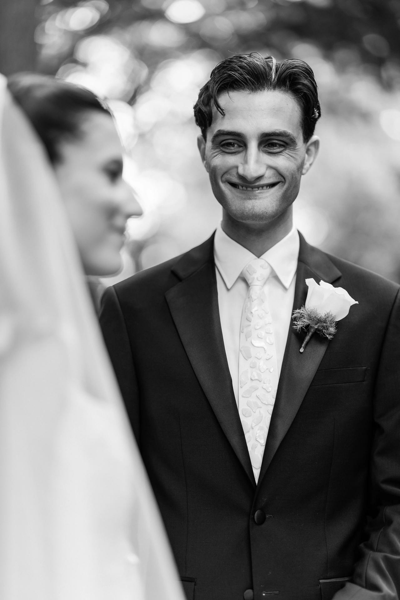 Black-and-white candid portrait of the groom smiling toward the camera while the bride stands close in the foreground, with soft bokeh lights in the background. Wedding photography at Crystal Plaza, Livingston, NJ.