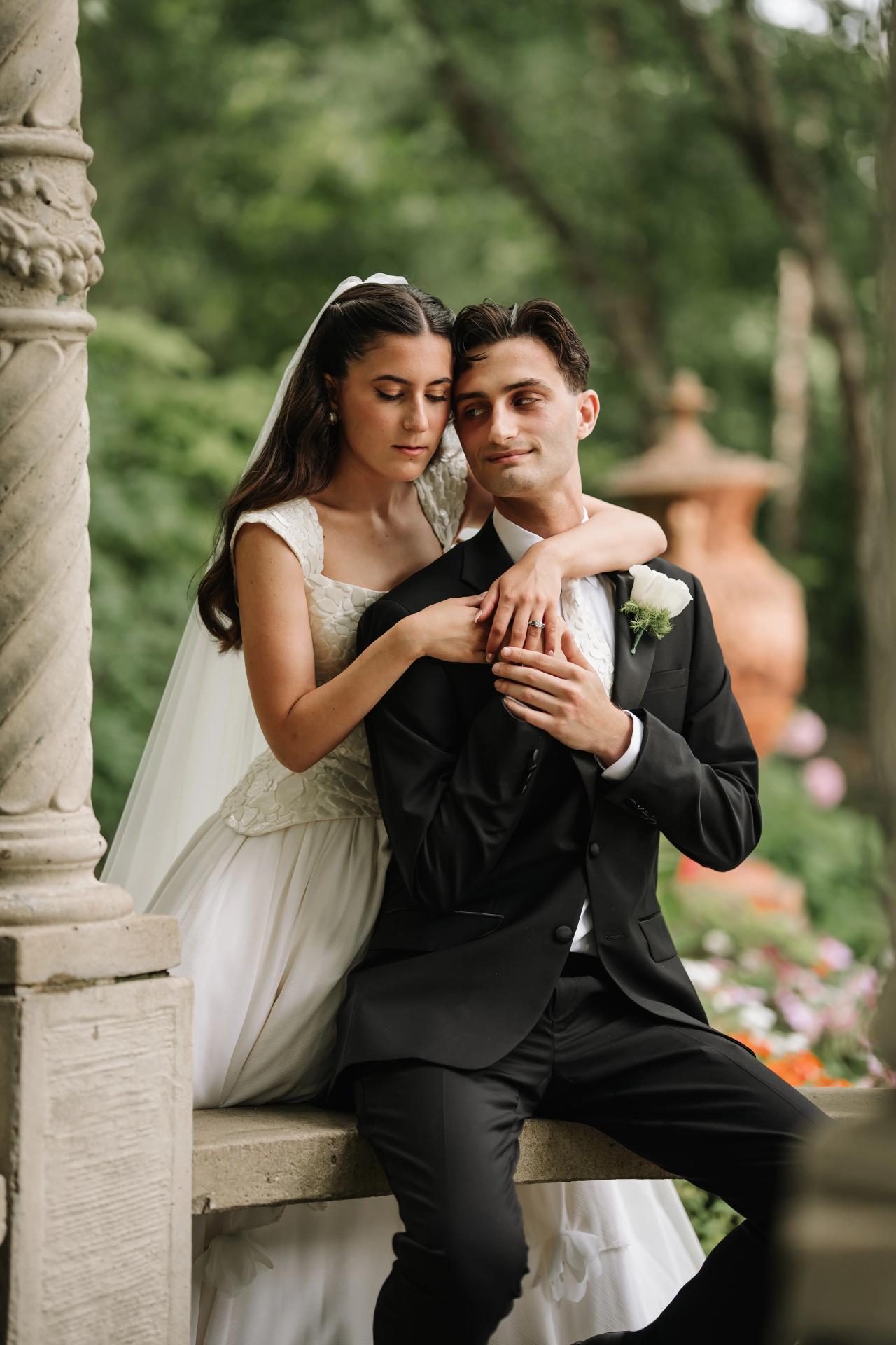 Bride and groom embracing tenderly while seated on stone steps in a lush garden, soft natural light and warm tones. Wedding photography at Crystal Plaza, Livingston, NJ.