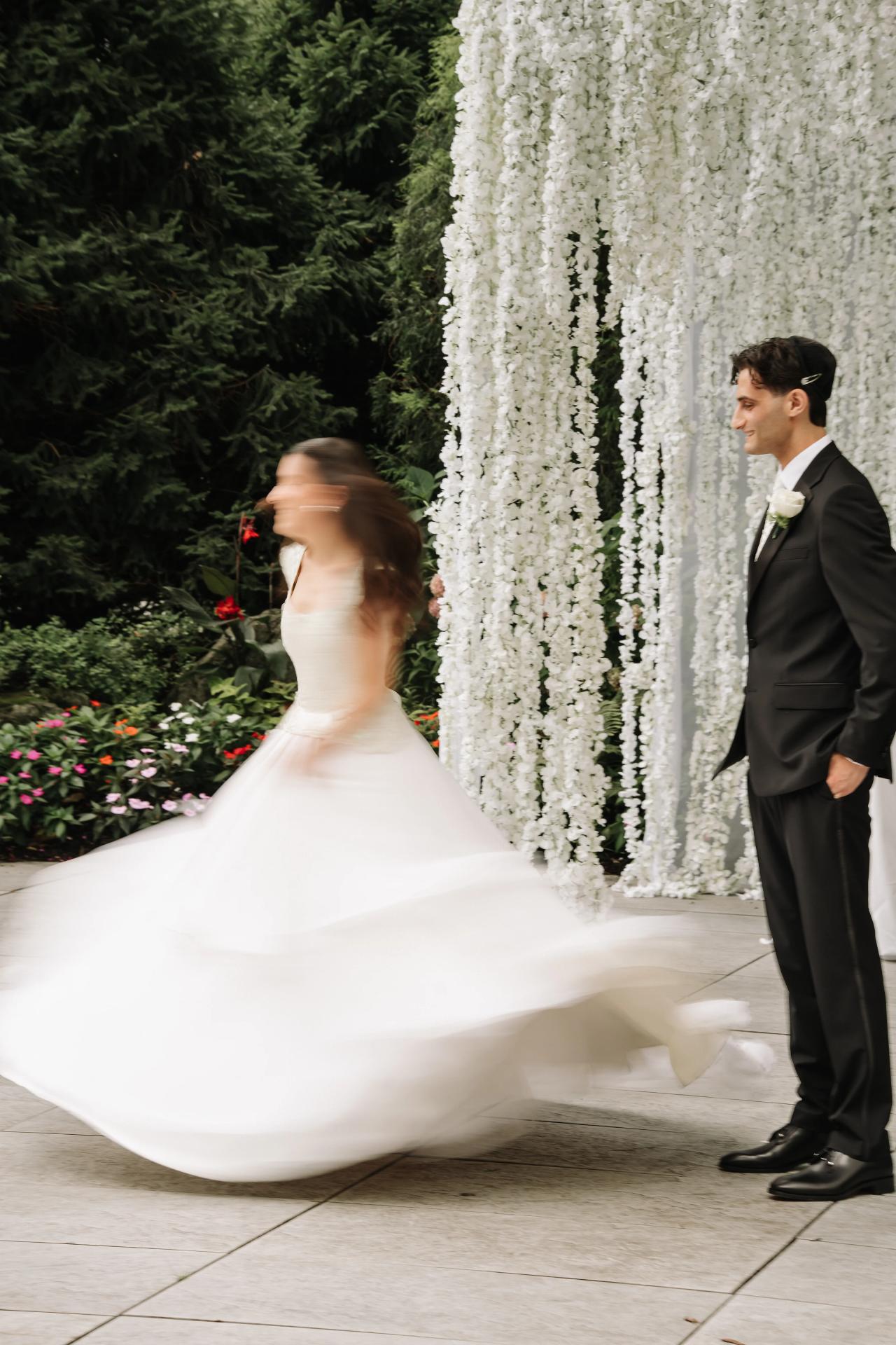 Bride in a flowing white gown and groom in a black suit stand near a tall white floral backdrop at a wedding chuppah, with the bride captured mid-motion. Wedding photography at Crystal Plaza, Livingston, NJ.