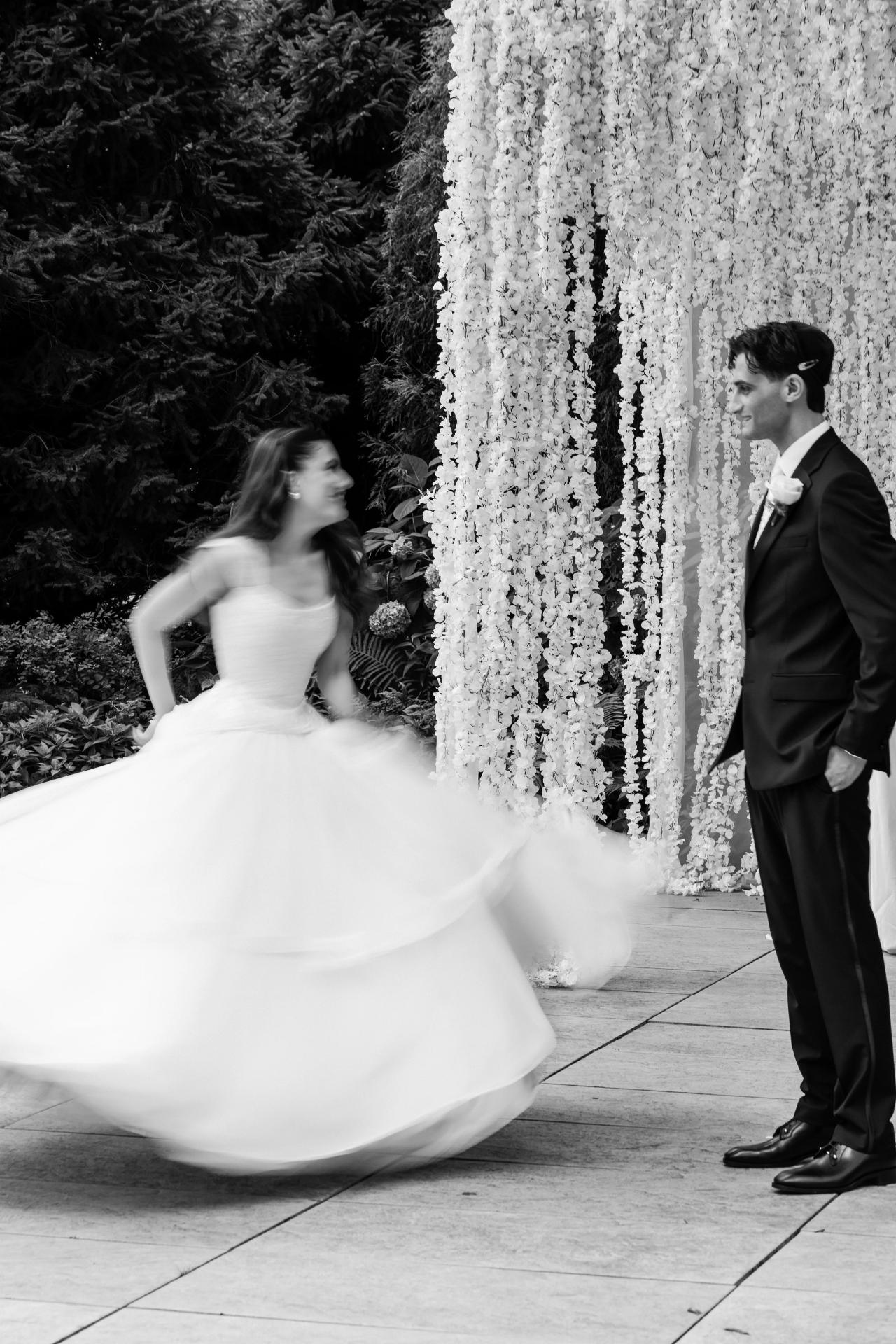 Black-and-white wedding photo of the bride and groom on a stone walkway beside a floral curtain backdrop, captured mid-spin with motion blur. Wedding photography at Crystal Plaza, Livingston, NJ.