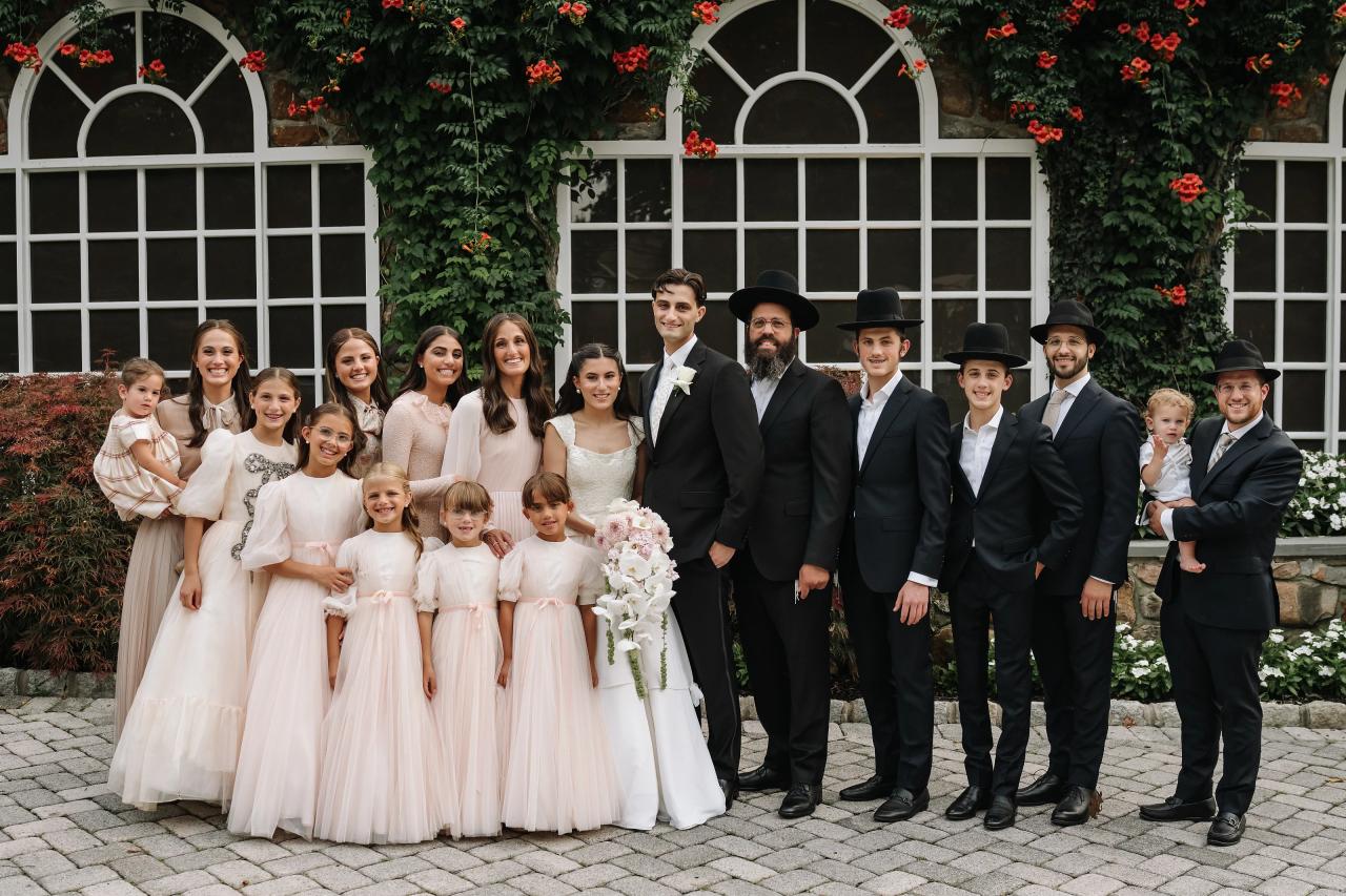Group wedding formal outdoors with bride and groom standing in front of arched greenery and red flowers, surrounded by family and children. Wedding photography at Crystal Plaza, Livingston, NJ.