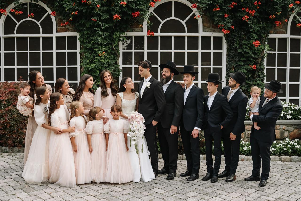Group of family members including a bride and groom posing outdoors in formal attire in front of a green wall with red flowers and arched windows. Wedding photography at Crystal Plaza, Livingston, NJ.