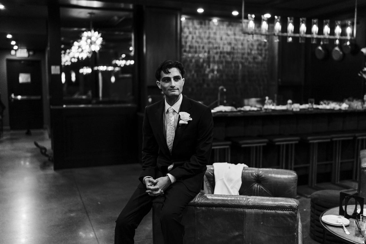 Black-and-white photo of the groom seated on a leather couch at a wedding reception, hands clasped, with the bar and chandeliers softly blurred in the background. Wedding photography at Crystal Plaza, Livingston, NJ.