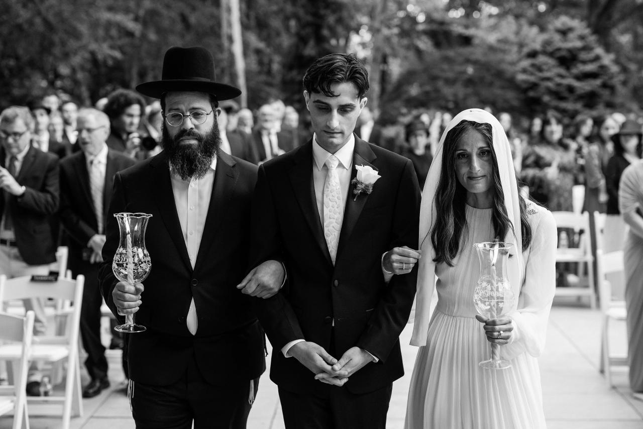 Black-and-white wedding portrait of a groom and bride in formal attire walking forward with close family members, holding decorative glasses in an indoor ceremony hall. Wedding photography at Crystal Plaza, Livingston, NJ.