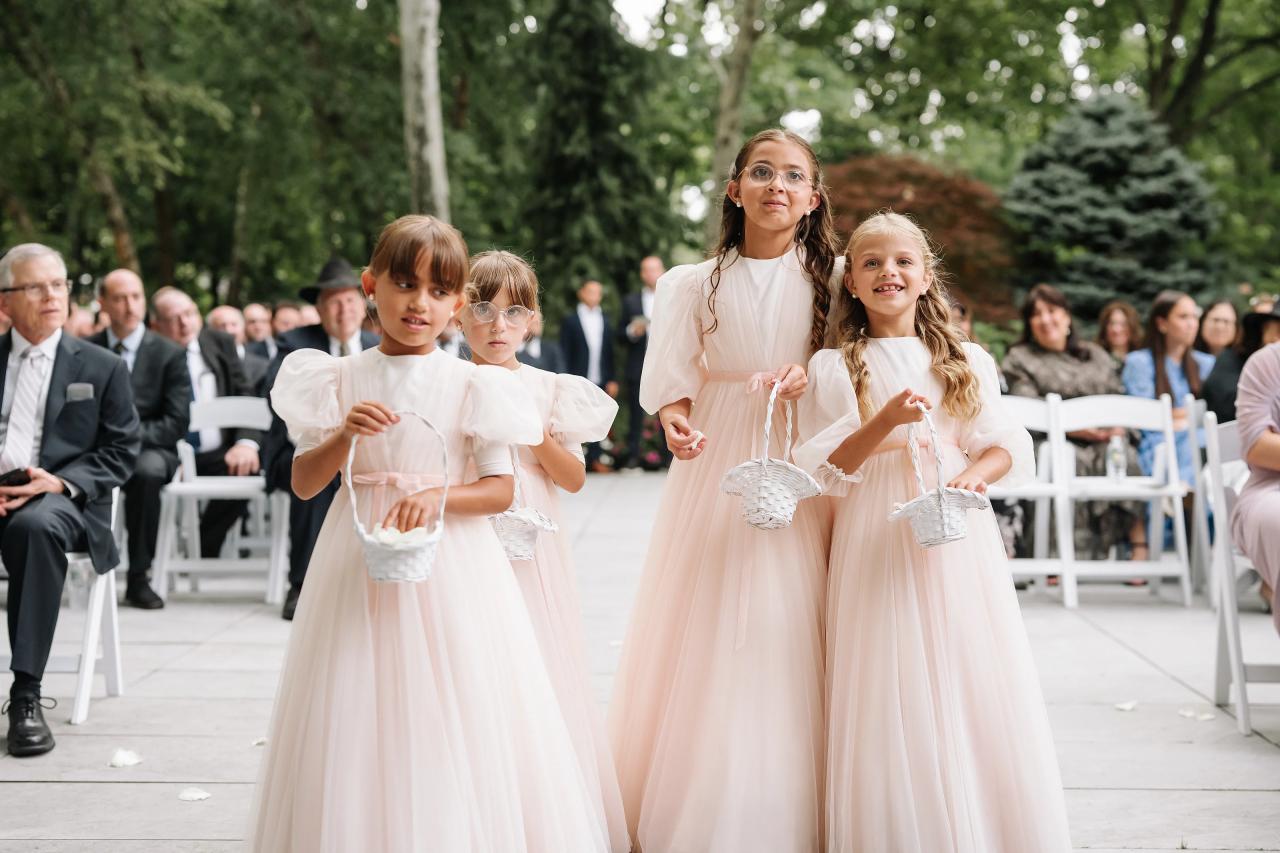 Bride in a white gown walking with two flower girls down an outdoor ceremony aisle, holding small baskets as they move toward the front. Wedding photography at Crystal Plaza, Livingston, NJ.