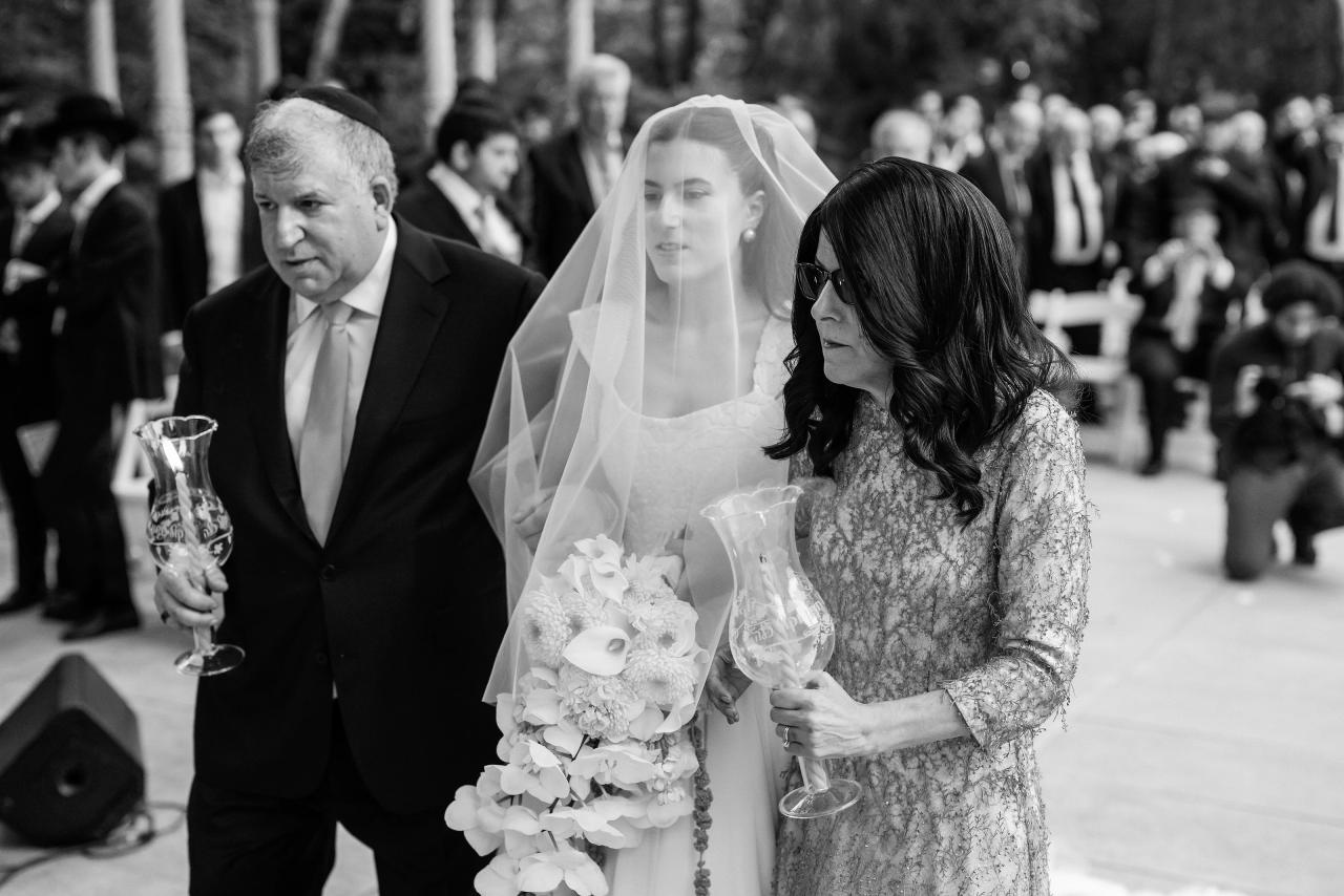 Black-and-white wedding ceremony moment: bride in veil holding a bouquet as an older woman stands close beside her, with a crowd of guests blurred in the background. Wedding photography at Crystal Plaza, Livingston, NJ.