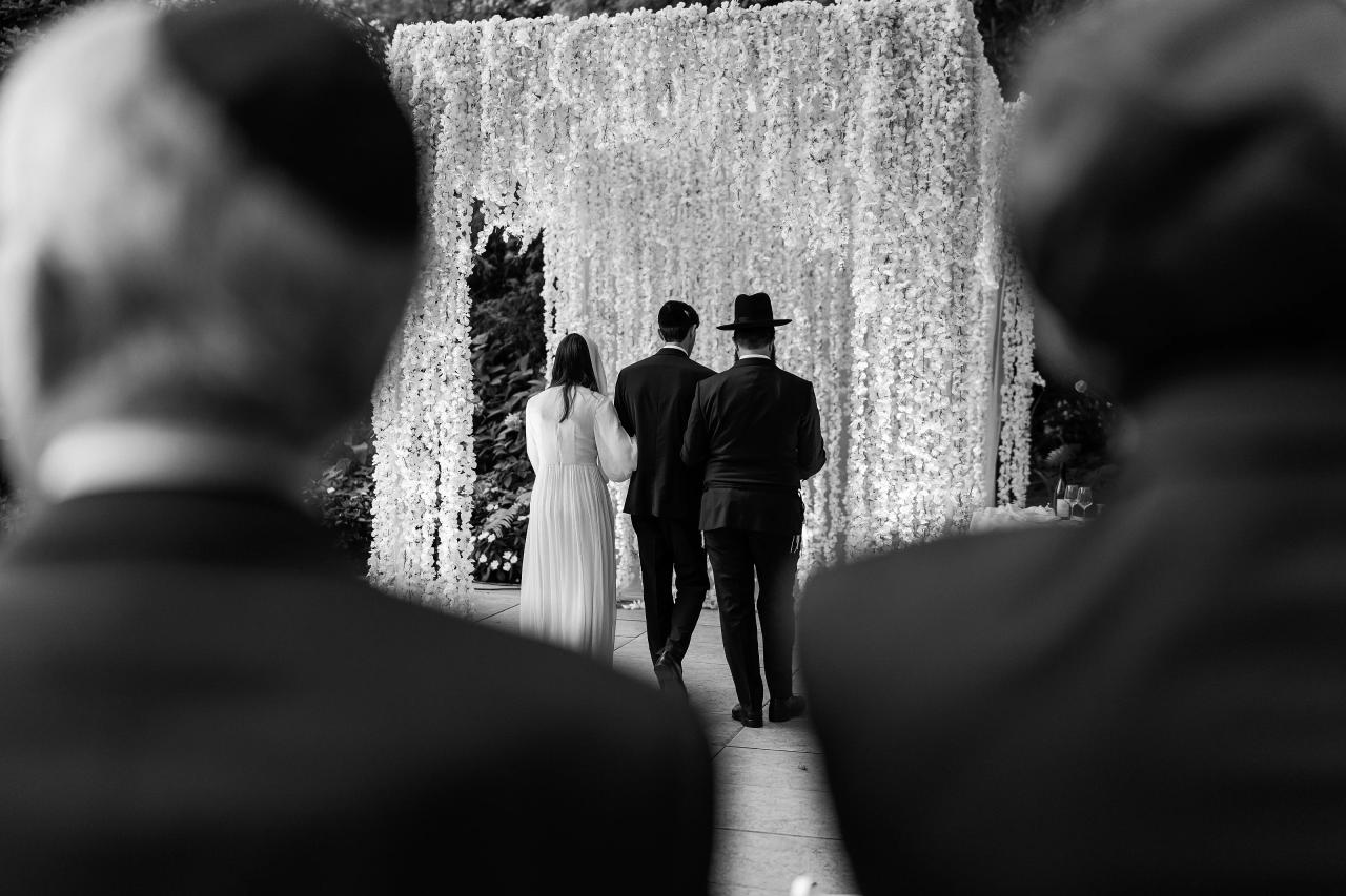 Black-and-white wedding photo showing the couple walking toward a flower-filled chuppah entrance down a lit aisle, framed by silhouettes in the foreground. Wedding photography at Crystal Plaza, Livingston, NJ.