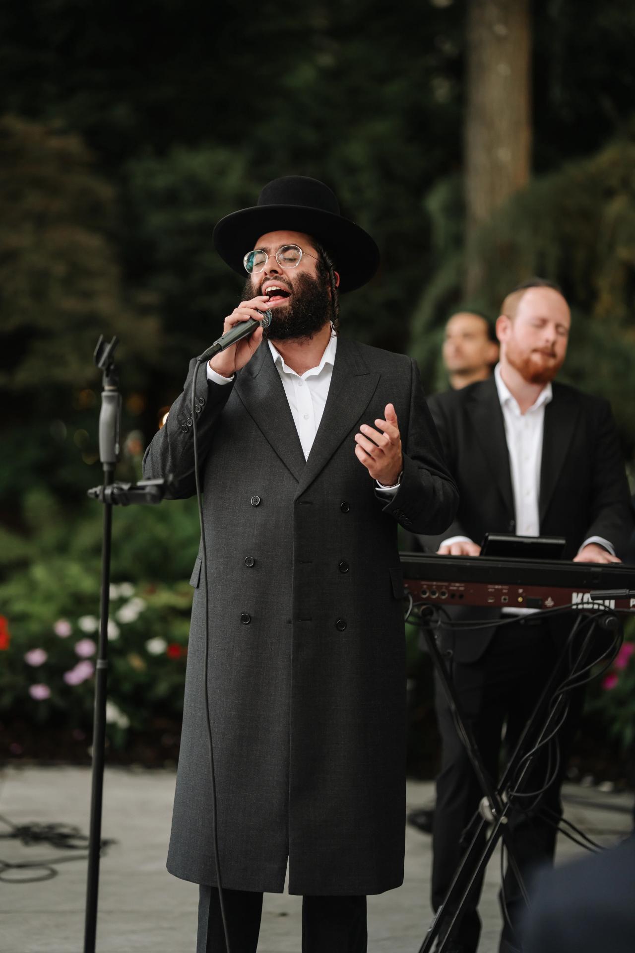 Outdoor event: a man in a dark traditional outfit with a black hat and glasses sings passionately into a microphone while a band plays behind him. Wedding photography at Crystal Plaza, Livingston, NJ.