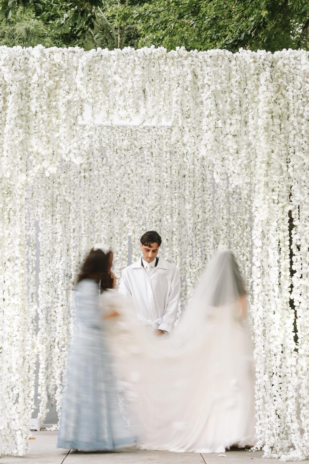Bride and groom framed by a white floral backdrop, with the bride blurred in motion while the groom stands in the center, heads bowed slightly. Wedding photography at Crystal Plaza, Livingston, NJ.