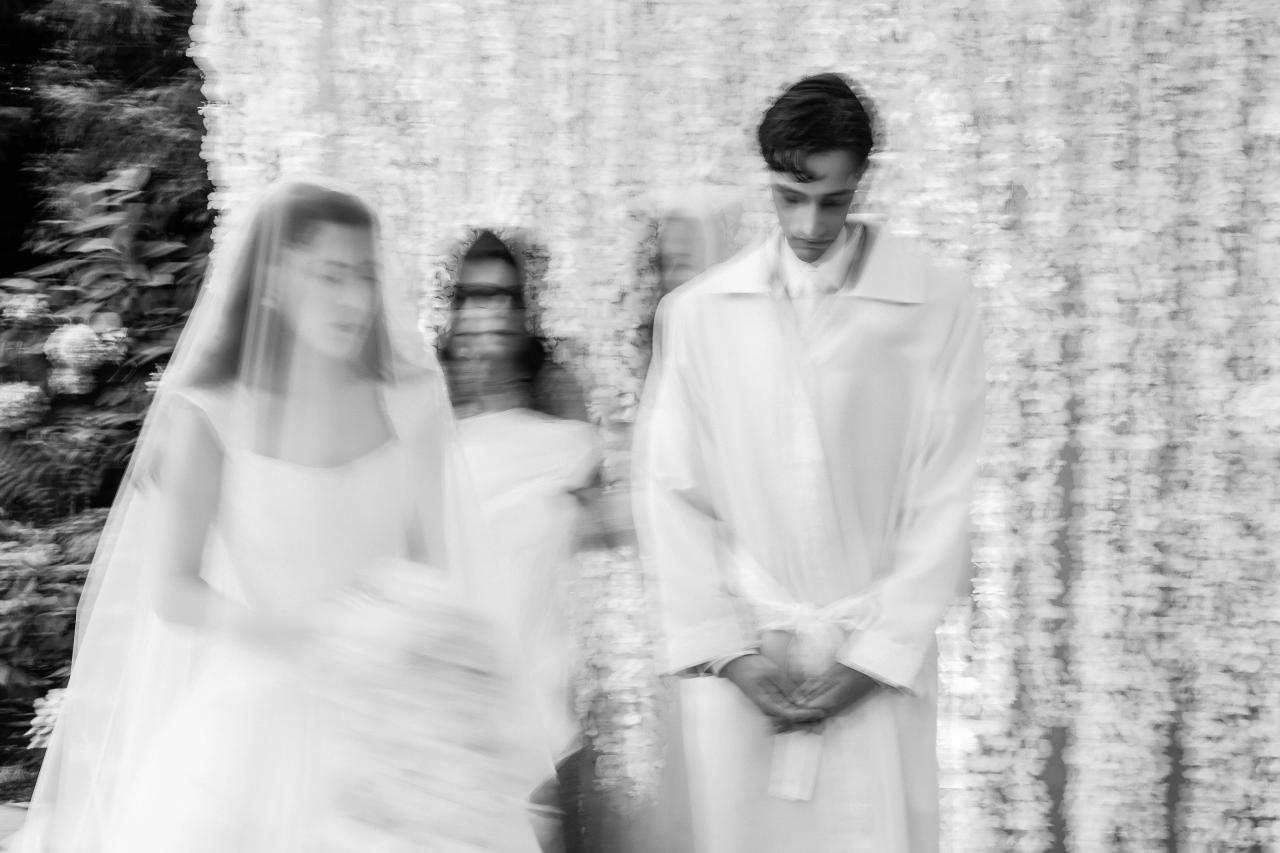 Black-and-white motion-blur photograph of a bride and groom walking side by side past a stone wall, with the bride’s veil creating a sweeping blur effect. Wedding photography at Crystal Plaza, Livingston, NJ.
