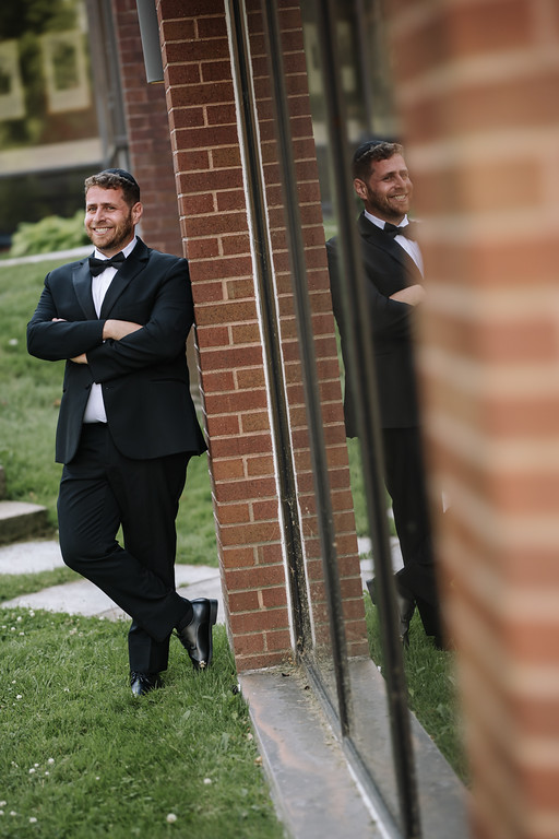 Two similar side-by-side views of a groom in a black tuxedo smiling outdoors near a brick wall and lawn, with soft background blur.