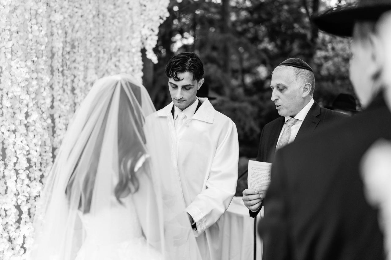 Black-and-white ceremony photo: a groom in a formal shirt stands beside an attendant reading ceremonial notes near floral drapery. Wedding photography at Crystal Plaza, Livingston, NJ.