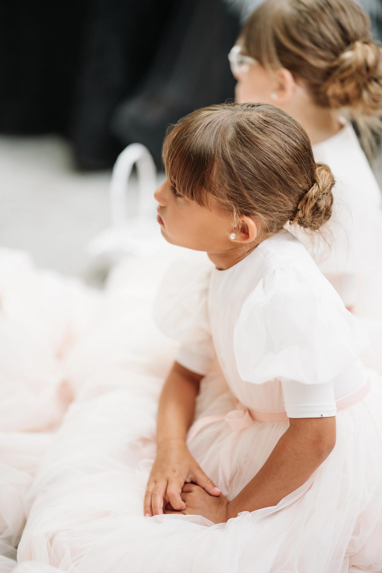 Framed side profile of two young girls in white dresses seated indoors, softly lit with blurred background. Wedding photography at Crystal Plaza, Livingston, NJ.
