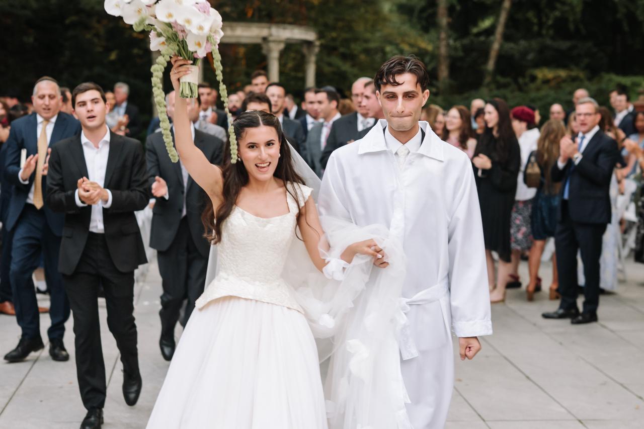 Bride and groom walking together in celebration outdoors, bouquet raised overhead while guests clap in the background. Wedding photography at Crystal Plaza, Livingston, NJ.