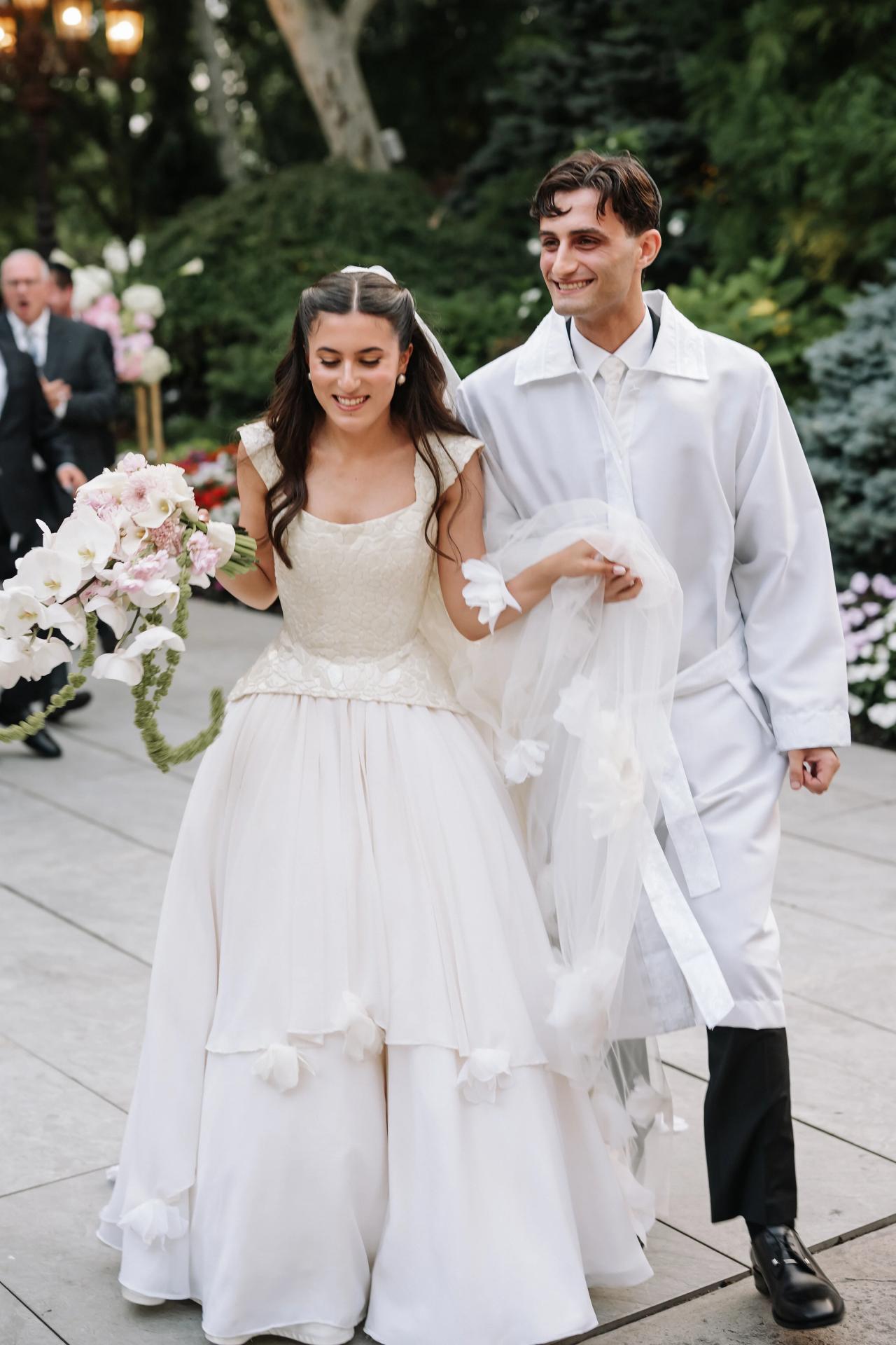 Framed wedding portrait of a bride and groom walking together down a stone walkway, the bride carrying a bouquet of white flowers and both smiling. Wedding photography at Crystal Plaza, Livingston, NJ.