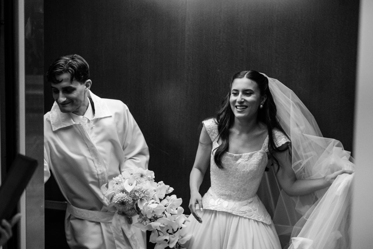 Black-and-white portrait of a bride and groom walking together indoors, smiling, with bouquets and the bride’s veil draped over her shoulder. Wedding photography at Crystal Plaza, Livingston, NJ.