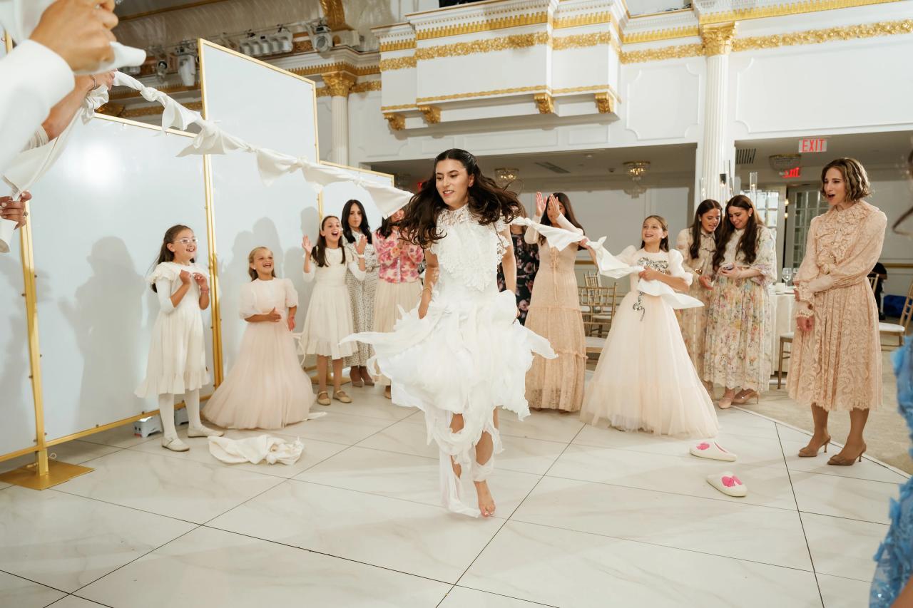 Bride dancing forward in a white dress in a bright venue hallway, with children and family members clapping and walking behind her; gold trim and an EXIT sign visible in the background. Wedding photography at Crystal Plaza, Livingston, NJ.