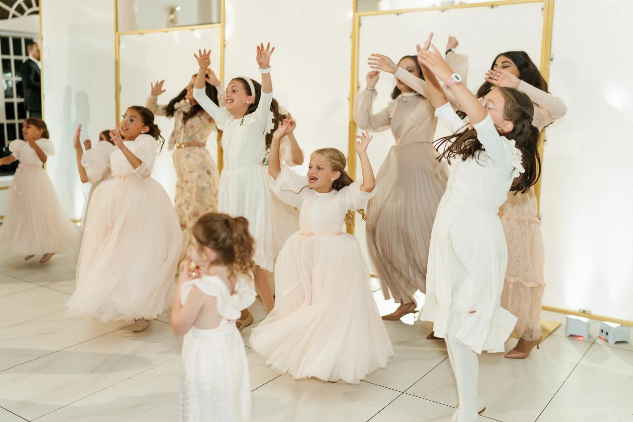Wide shot of joyful guests and children dancing with arms raised in a bright white indoor hall. Wedding photography at Crystal Plaza, Livingston, NJ.