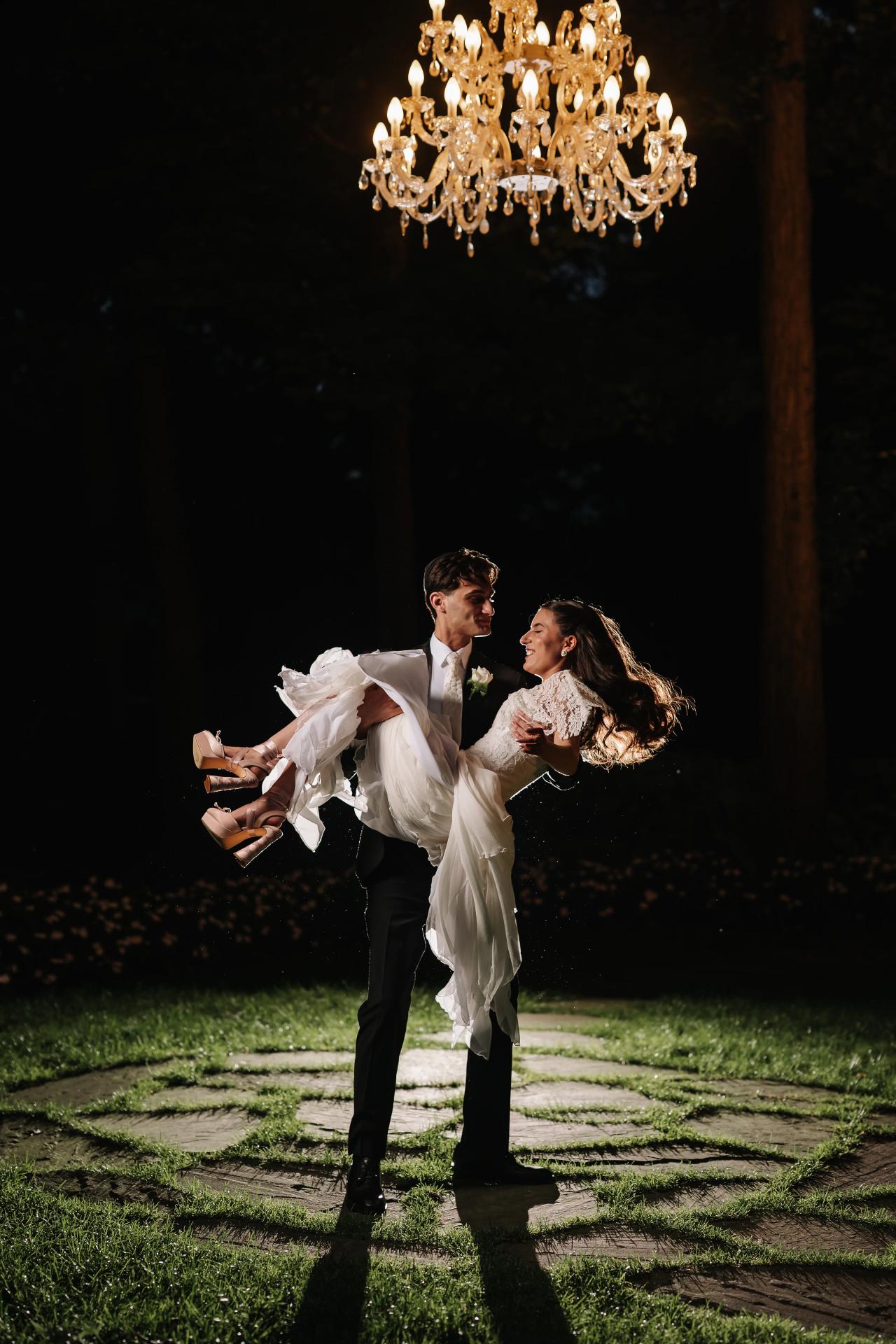 Bride and groom outdoors at night jumping mid-moment under a crystal chandelier, laughing, with bright spot of lawn and stone path beneath them. Wedding photography at Crystal Plaza, Livingston, NJ.