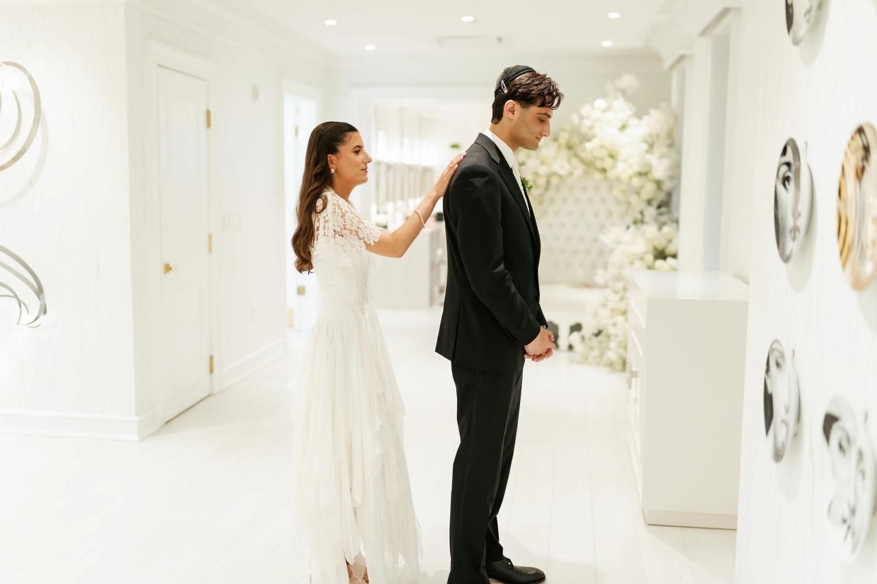 Bride and groom walking hand-in-hand in a bright white hallway, bride’s hand resting on groom’s shoulder, soft indoor lighting and framed decor in background. Wedding photography at Crystal Plaza, Livingston, NJ.