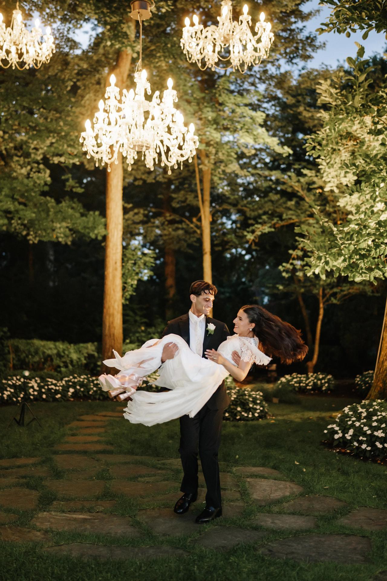 Bride and groom dancing in a warm evening garden under glowing chandelier lights, with the bride’s veil and dress lifted mid-spin. Wedding photography at Crystal Plaza, Livingston, NJ.