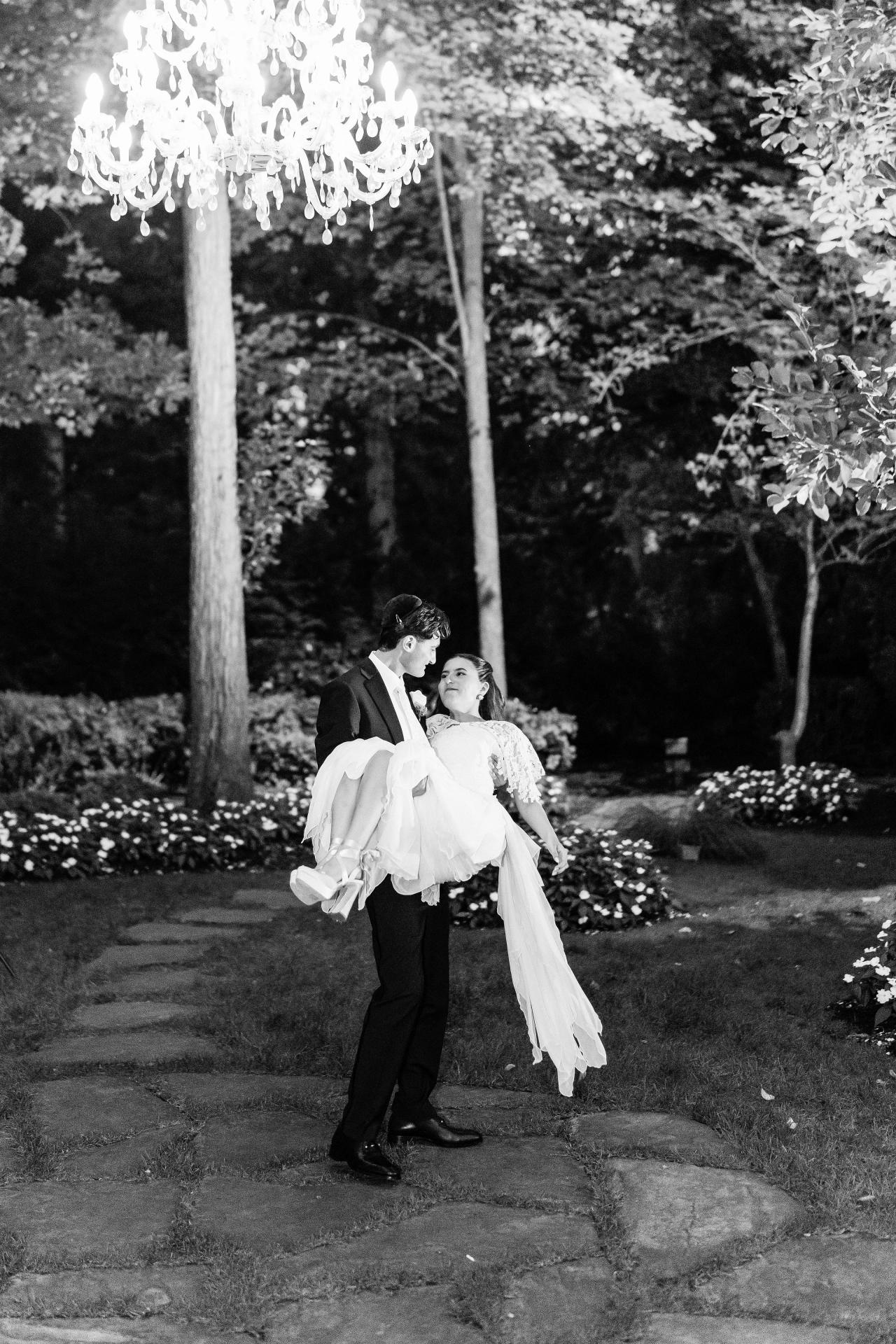 Black-and-white image of a bride and groom walking closely together at night in a garden under chandelier lights, with the bride’s veil flowing. Wedding photography at Crystal Plaza, Livingston, NJ.