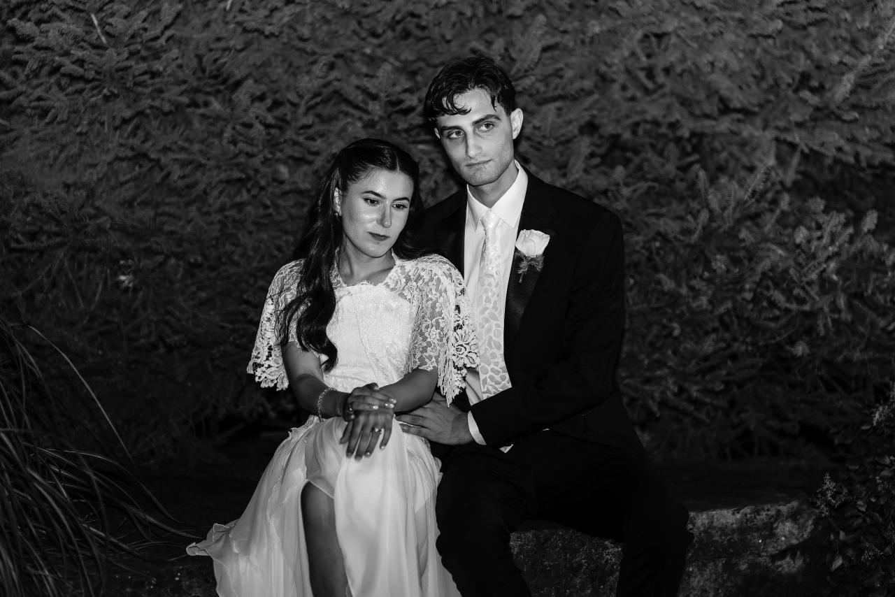 Black-and-white portrait of a Jewish bride and groom in formal attire sitting close together outdoors, with the bride seated and groom kneeling behind her. Wedding photography at Crystal Plaza, Livingston, NJ.