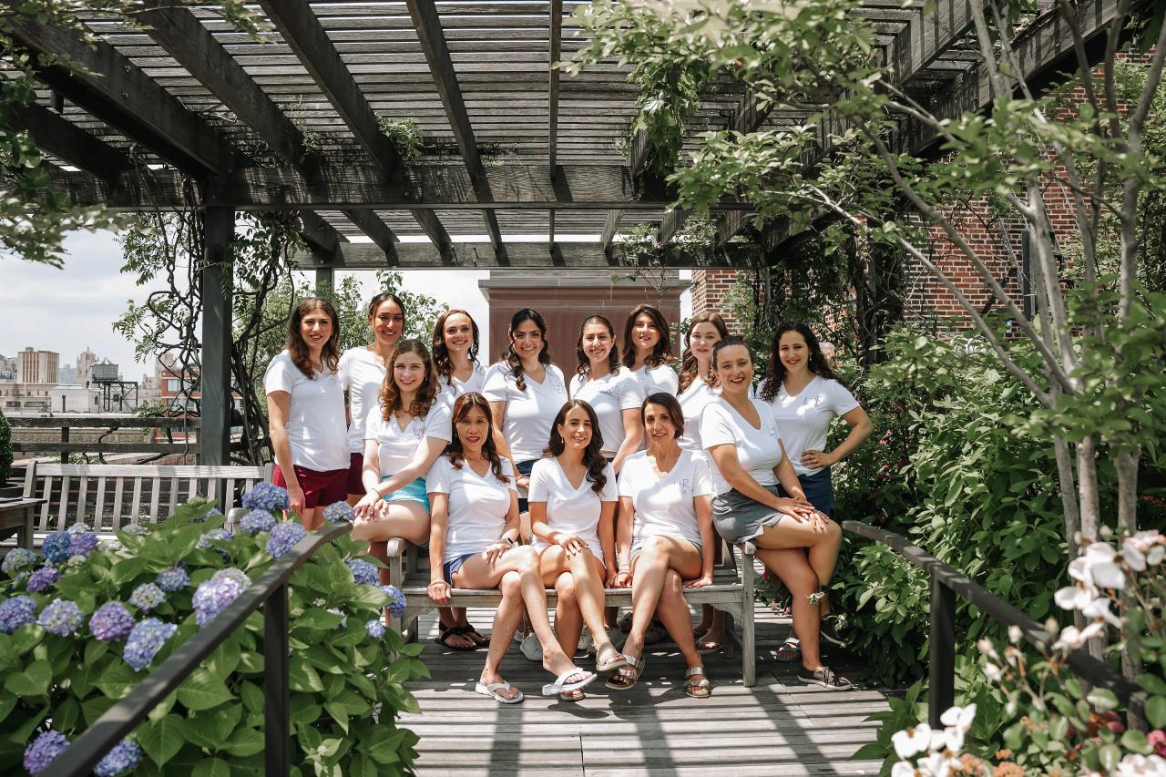 A posed group of women in white dresses sitting on a wooden bench in a garden courtyard under a pergola, surrounded by green leaves and blue flowers. Wedding photography at Tribeca Rooftop + 360°, New York, NY.