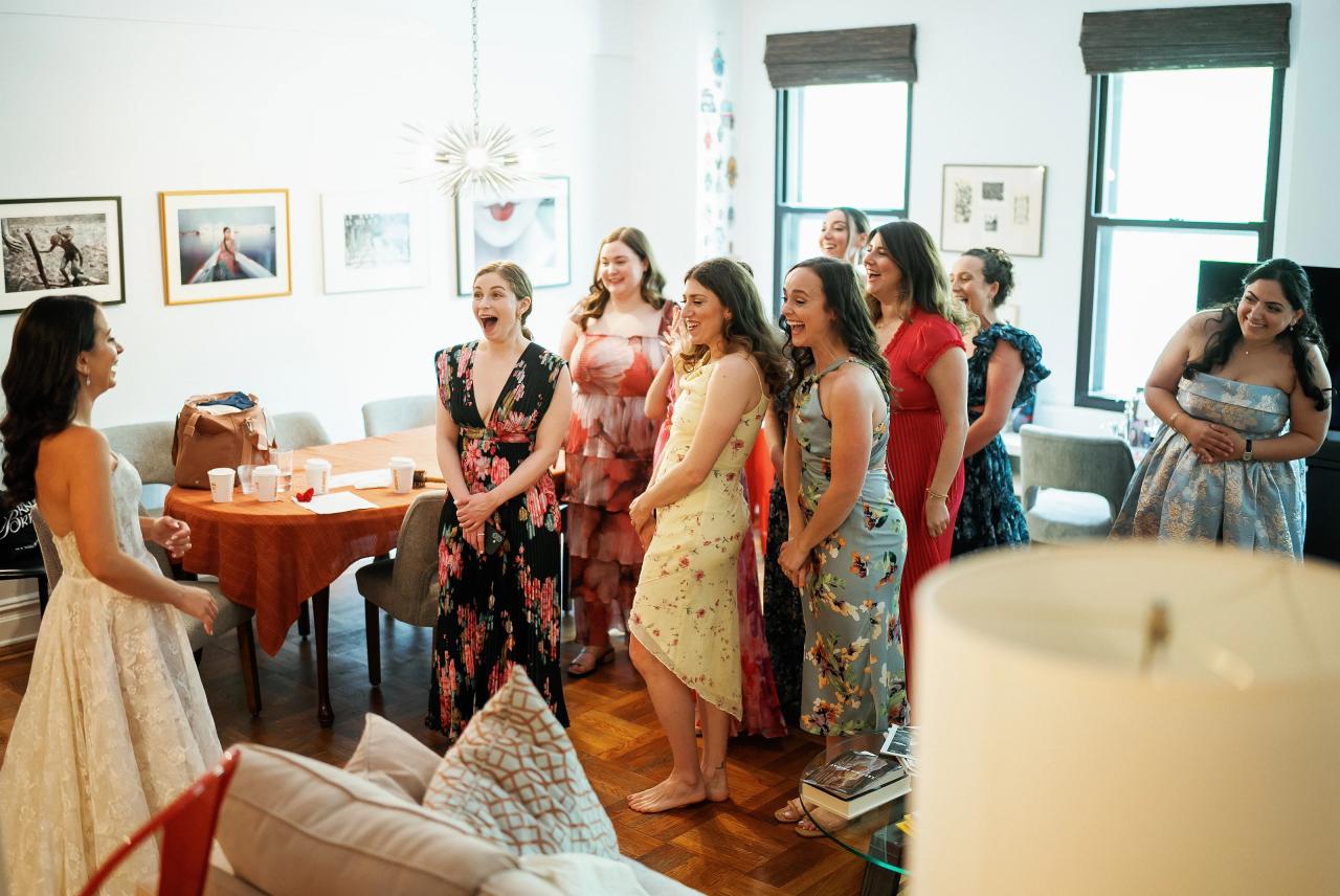Group of guests standing and smiling inside a bright reception room with windows and framed wall art, watching something off-camera. Wedding photography at Tribeca Rooftop + 360°, New York, NY.
