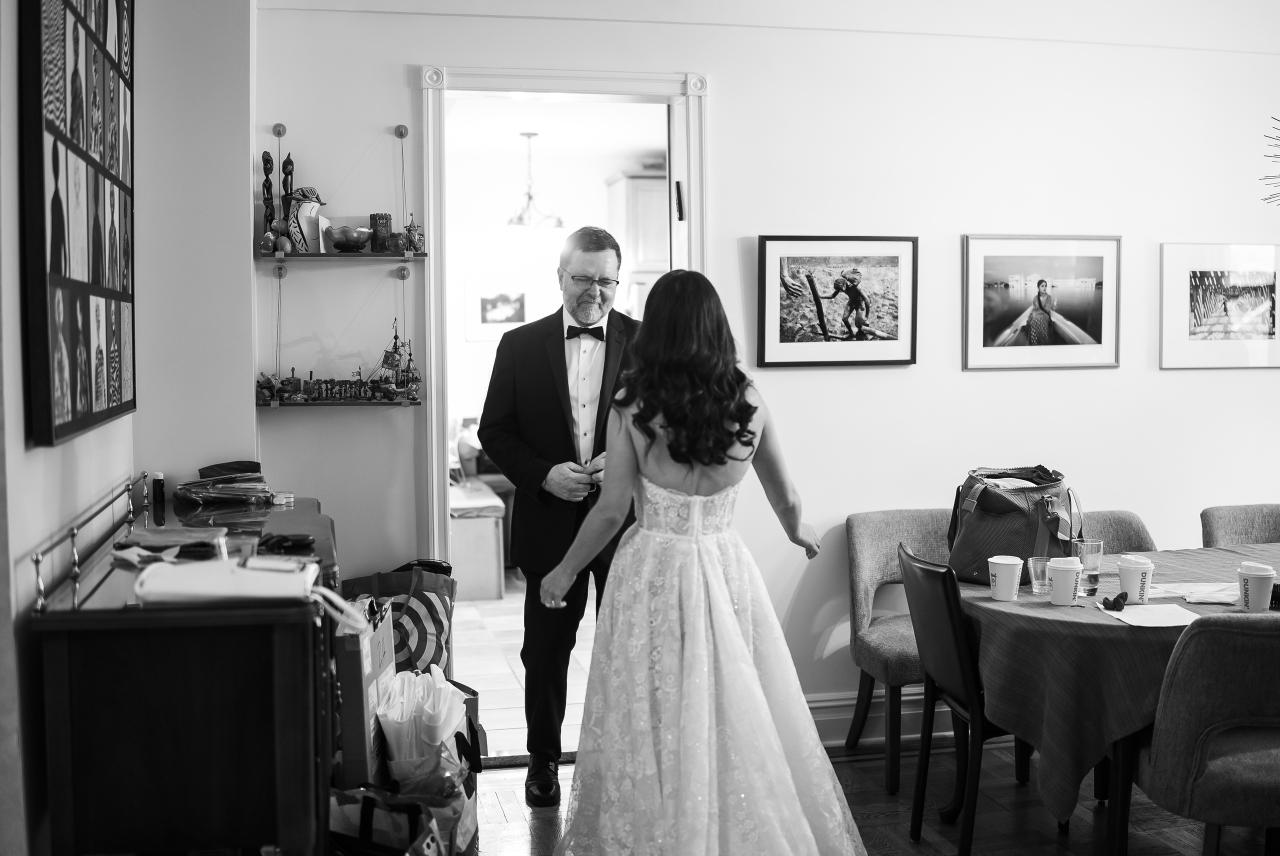 Black-and-white photo of a bride in a light gown walking with a suited man in a getting-ready room, with bright doorway light behind them. Wedding photography at Tribeca Rooftop + 360°, New York, NY.