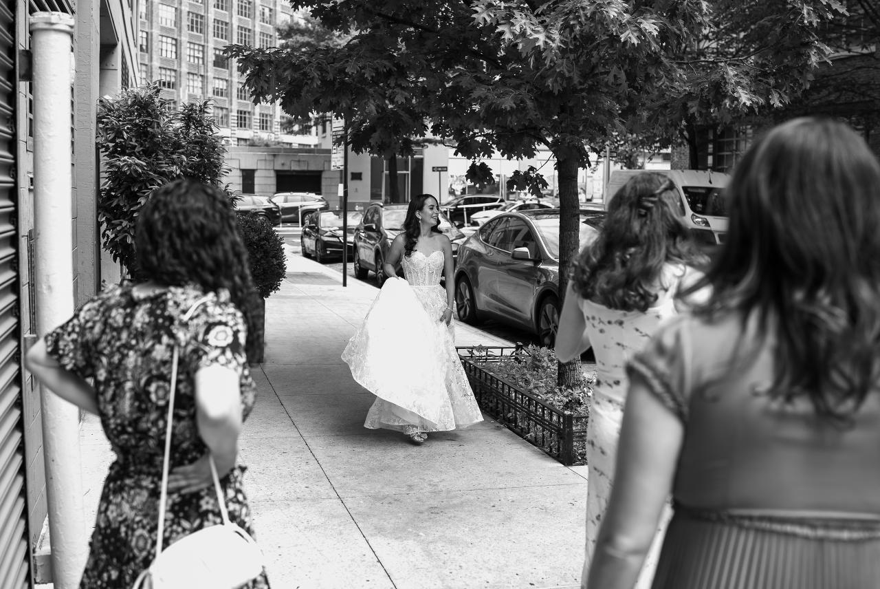 Black-and-white street portrait of a bride walking toward the camera, framed by columns and trees, with two guests in the foreground and parked cars in the background. Wedding photography at Tribeca Rooftop + 360°, New York, NY.