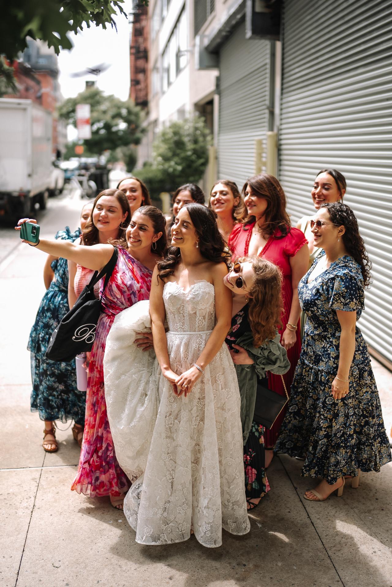 Bride walking outdoors with a group of smiling friends and family in colorful dresses; one guest raises a phone to film. Wedding photography at Tribeca Rooftop + 360°, New York, NY.