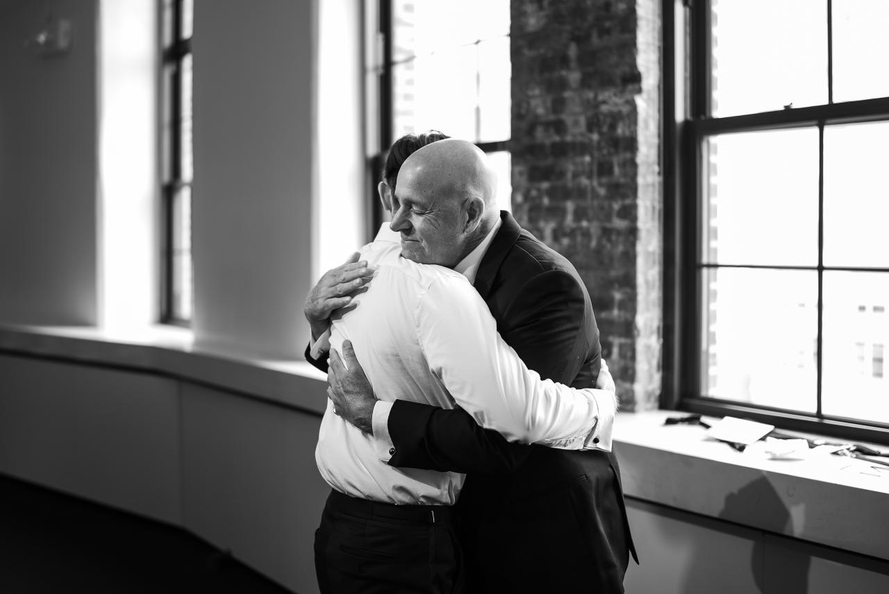 Black-and-white wedding photo of a joyful groom hugging an older man in a warm embrace by large window light. Wedding photography at Tribeca Rooftop + 360°, New York, NY.