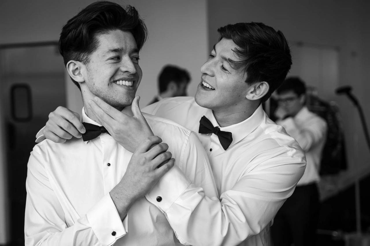 Black-and-white close-up of two men in dress shirts and bow ties laughing and adjusting each other’s bow ties during a wedding celebration, with softly blurred guests in the background. Wedding photography at Tribeca Rooftop + 360°, New York, NY.